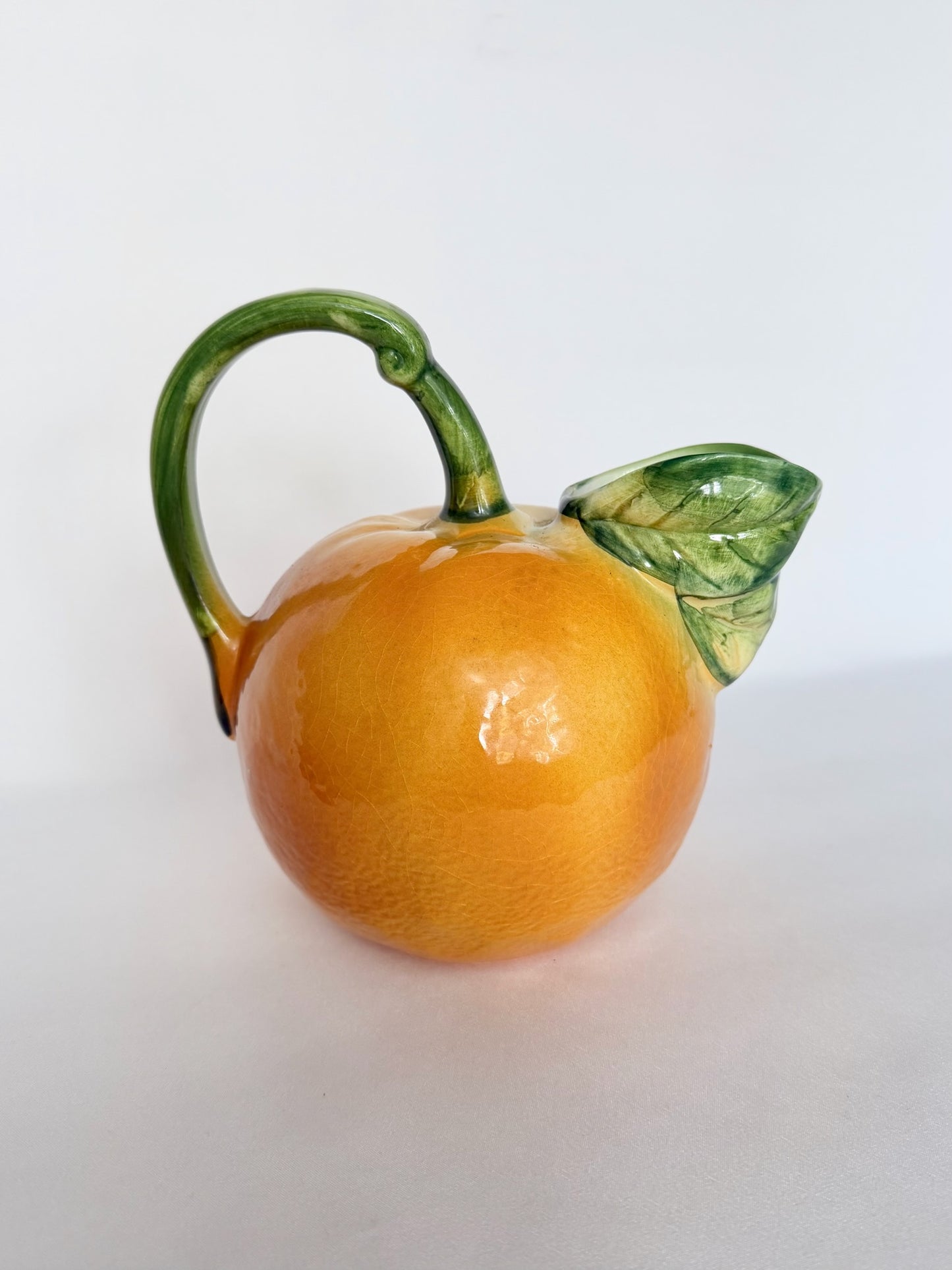 A vintage glazed ceramic pitcher shaped like an orange, featuring a vibrant orange body, a green stem forming the handle, and a detailed green leaf on the side, made in Italy