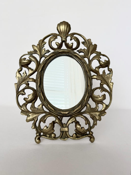 Vintage brass table top mirror with swirly acanthus leaves design and kick stand in back.
