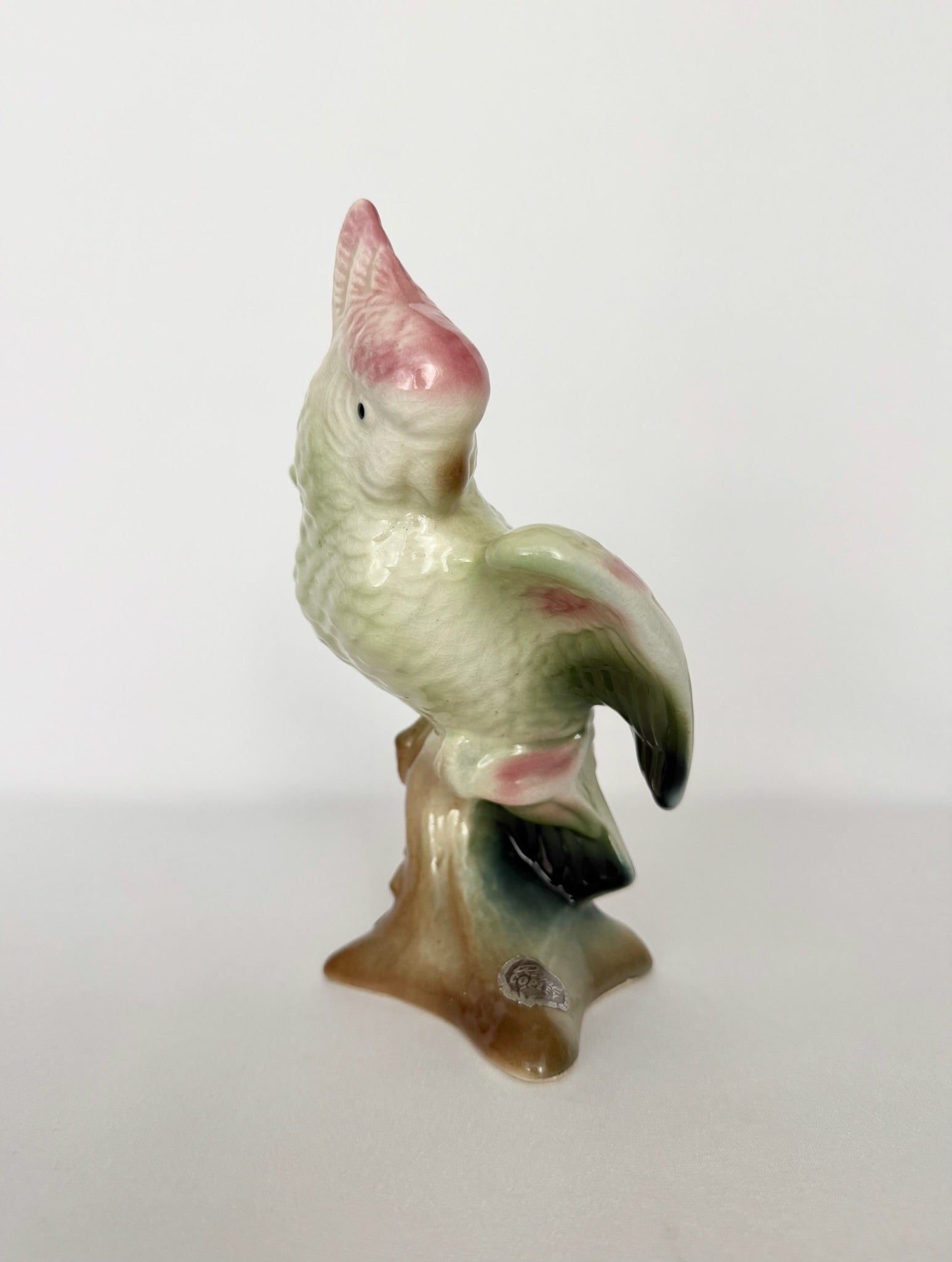 A vintage ceramic cockatoo parrot figurine perched on a small base, featuring a glossy glaze with pink, green, and cream colors.