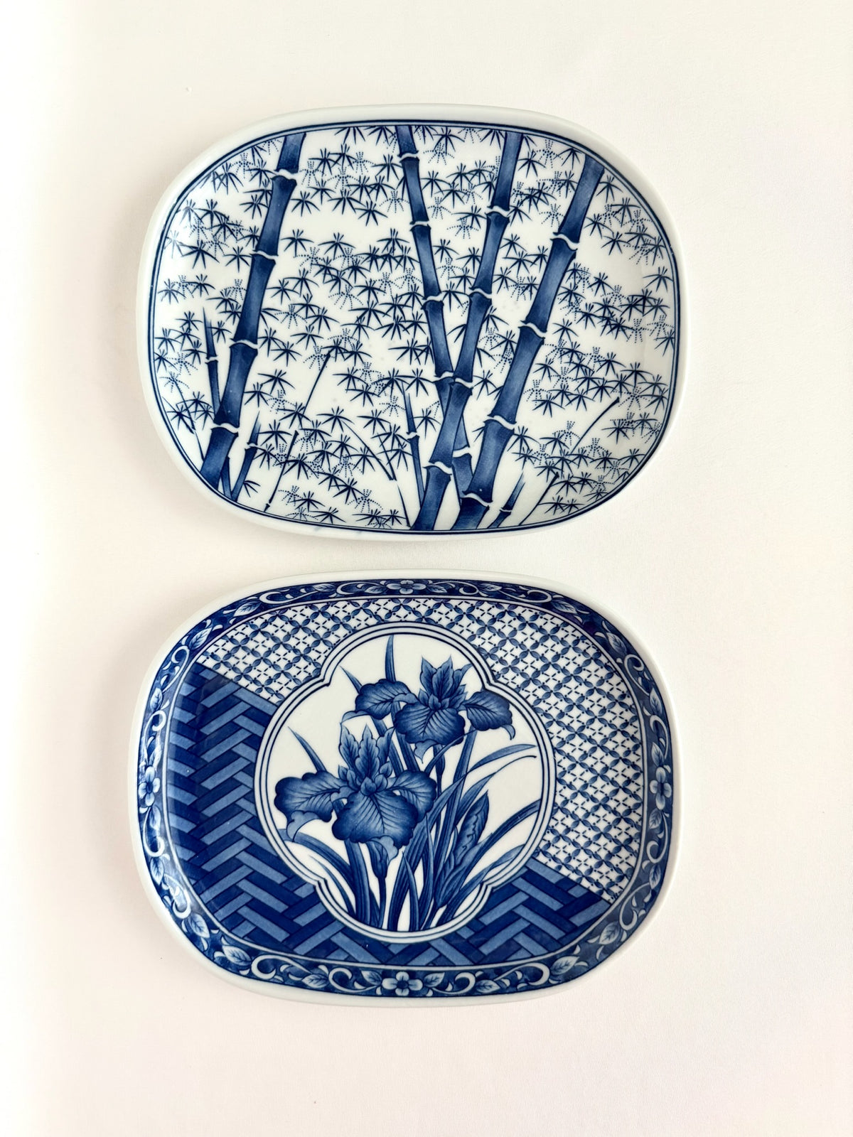 Two blue and white porcelain chinoiserie style plates with rounded rectangular shapes, stacked vertically. The top plate features a bamboo pattern, and the bottom plate features a floral iris pattern with geometric borders.