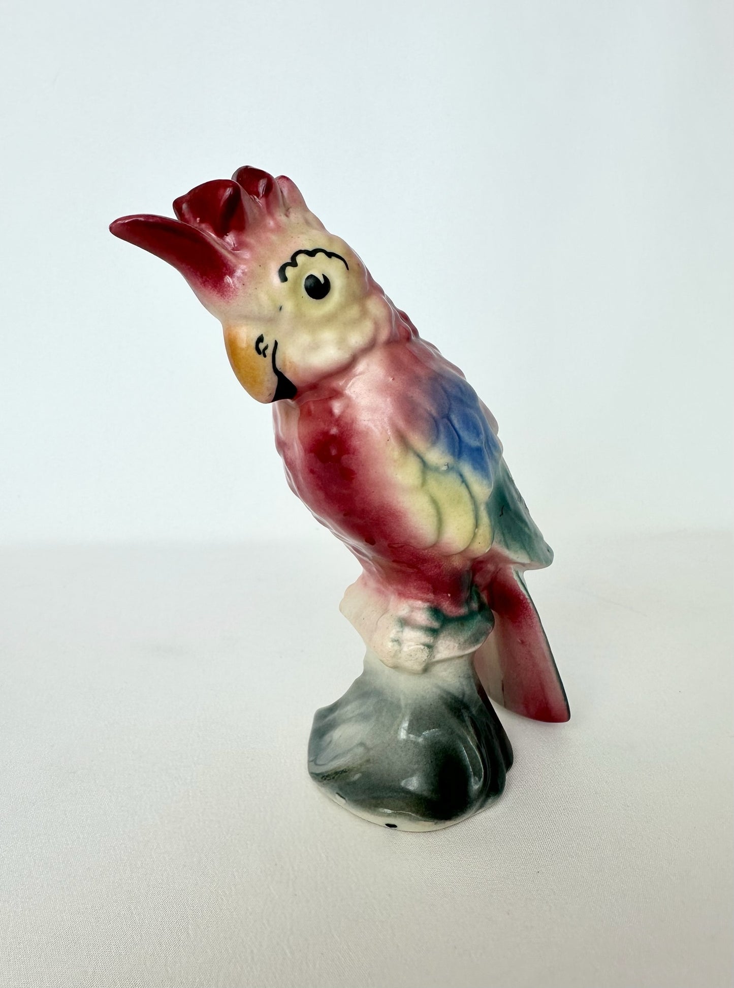 A vintage ceramic parrot figurine standing on a small base, featuring a red crest, cream face, yellow beak, and multicolored body with red, blue, green, and cream tones.