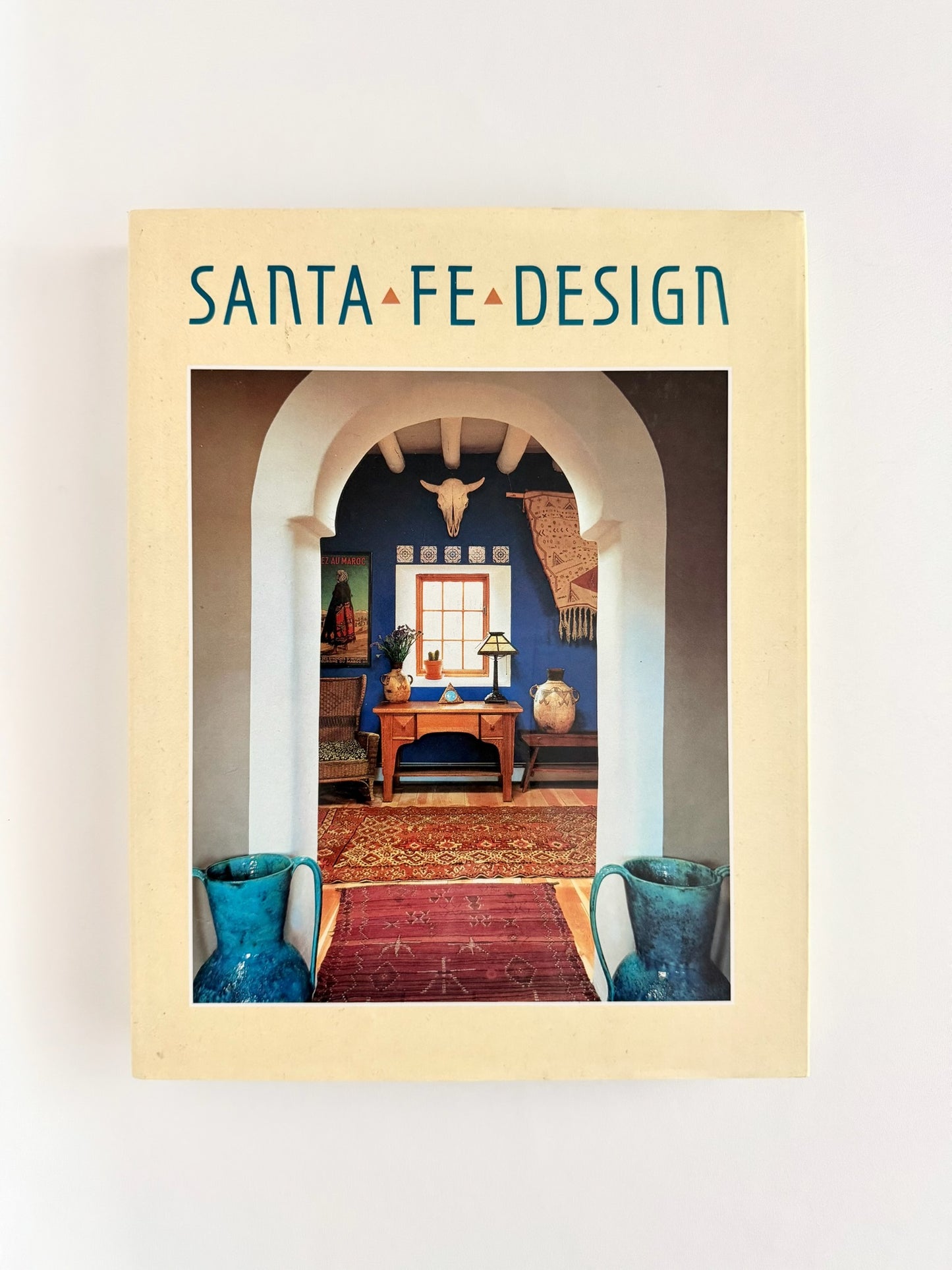 A vintage book titled 'SANTA FE DESIGN' with a cream-colored cover, featuring a central photograph of an arched interior doorway with blue walls, a wooden table, and a red patterned rug.