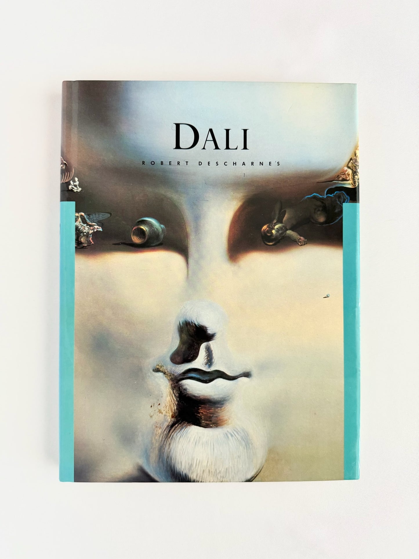 A vintage hardcover book titled 'DALI' by Robert Descharnes, featuring a surreal face illustration on the cover with turquoise side borders